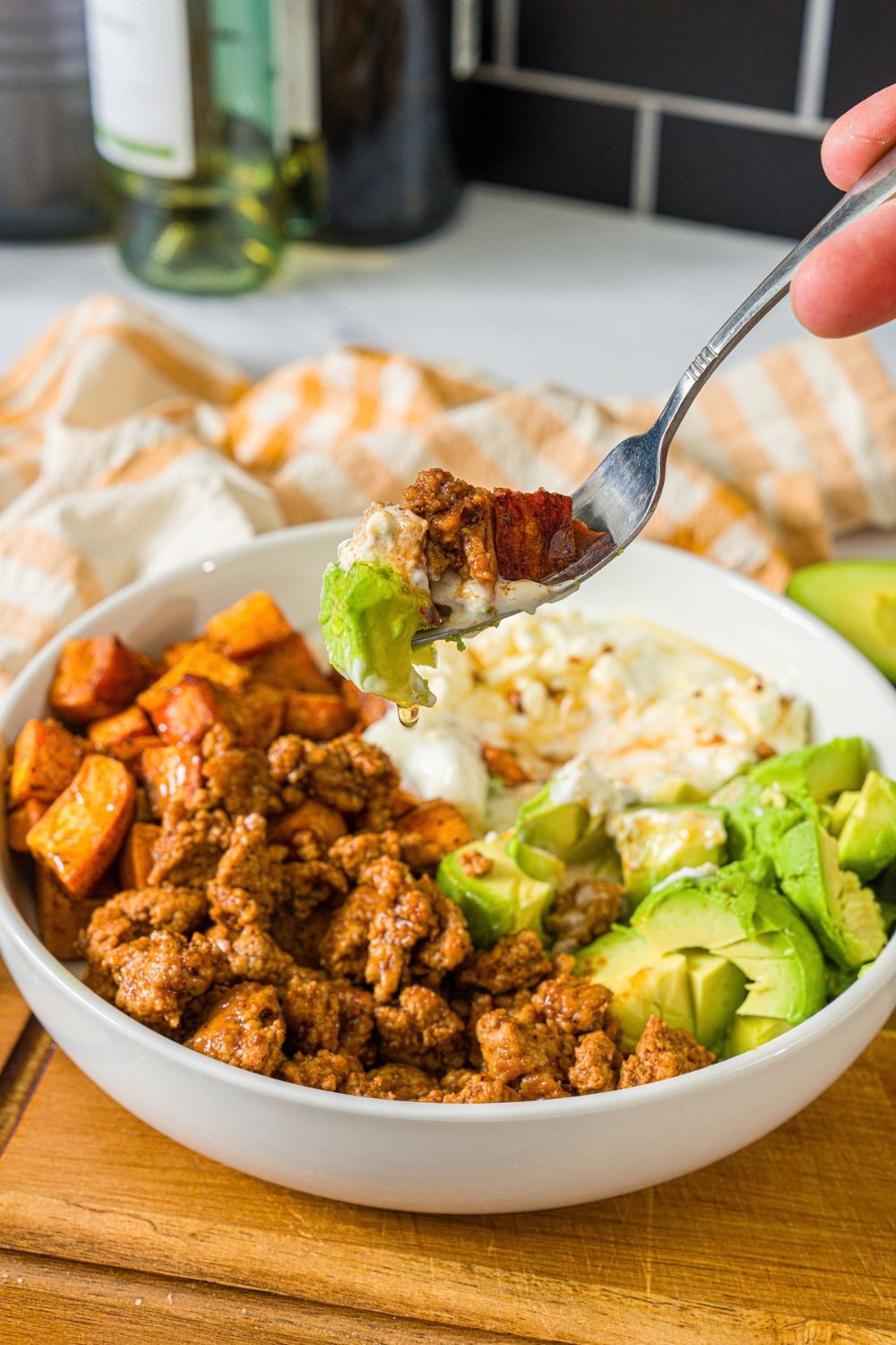 A white bowl with seasoned ground turkey, roasted sweet potatoes, avocado, and greek yogurt topped with hot honey. A fork is taking a bite from the bowl. The bowl is on a wooden board with a yellow checkered napkin.