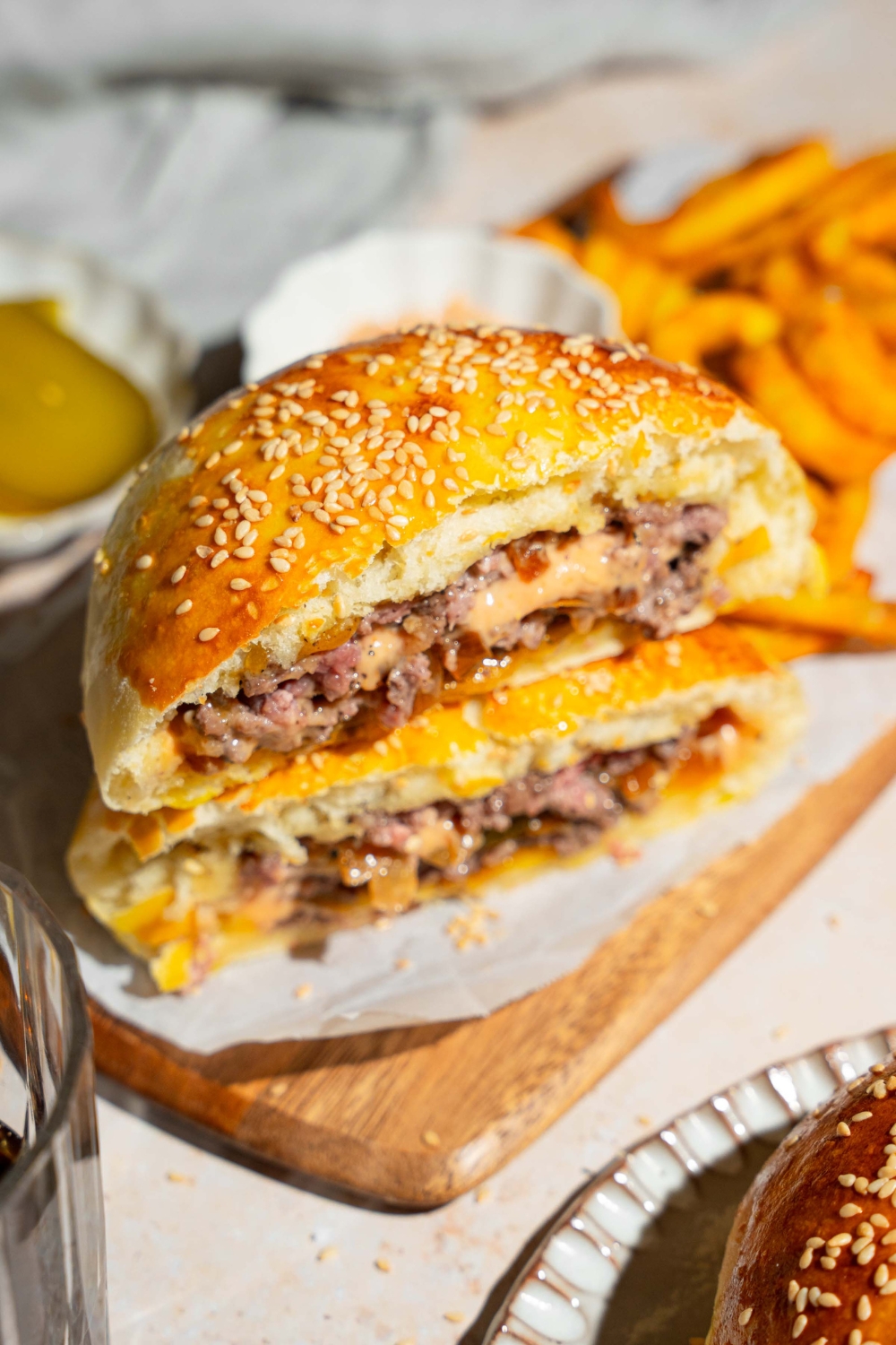 A wooden board lined with parchment paper with a burger stuffed bun sliced in half and stacked on top of each other. There are curly fries on the board.