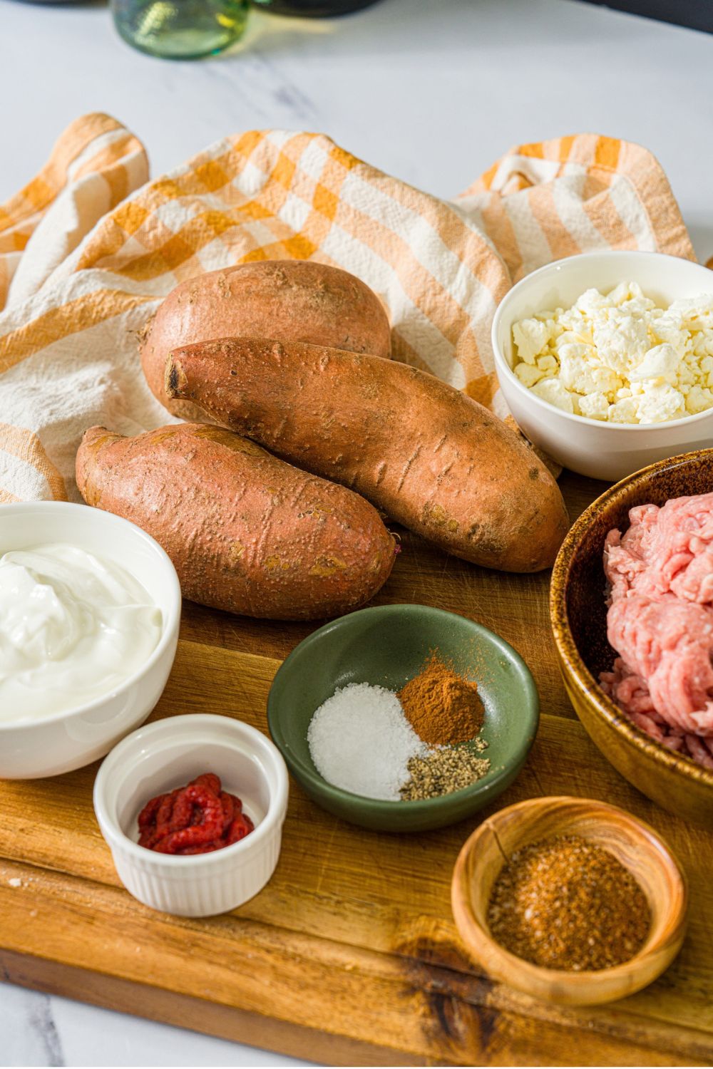 A wooden board with ingredients to make hot honey ground turkey sweet potato bowls including sweet potatoes, ground turkey, yogurt, tomato paste, and seasonings.
