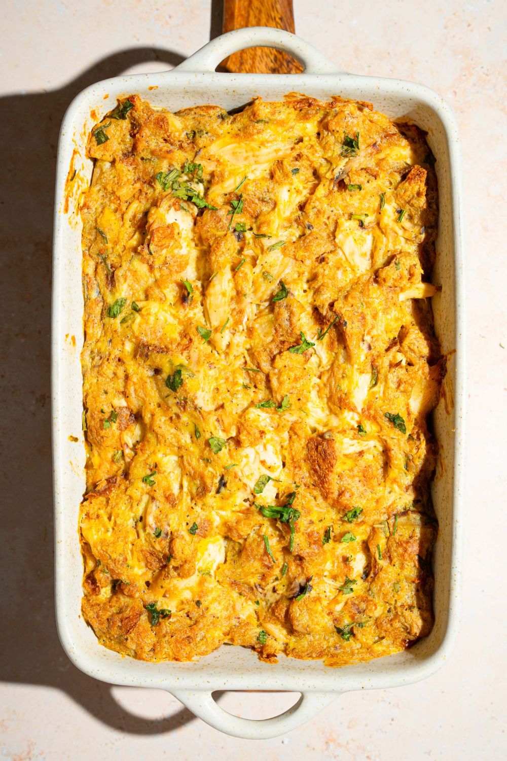 A baking dish with Dolly Parton's chicken and stuffing casserole garnished with fresh parsley. The dish s on a wooden board on a tan counter.