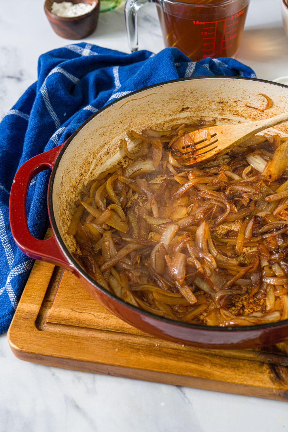A dutch oven with a spoon stirring caramelized onions. The dutch oven is on a wooden board with a blue cloth napkin.