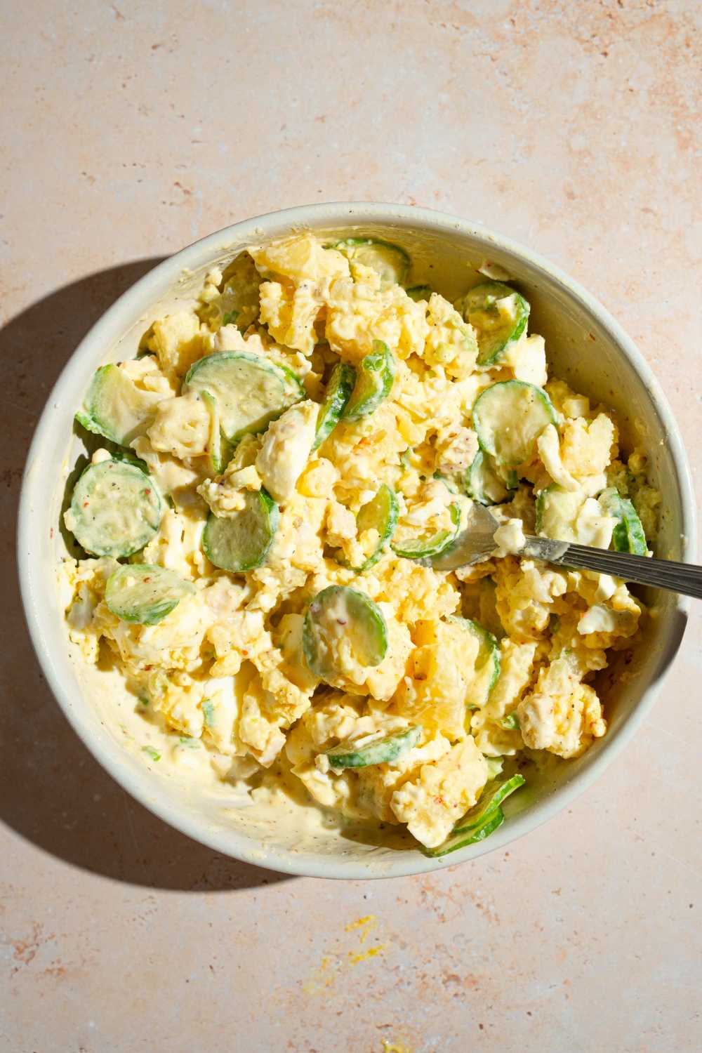 A white bowl with Japanese potato salad mixture with egg, chicken, cucumber, and sliced green onion in a creamy dressing.