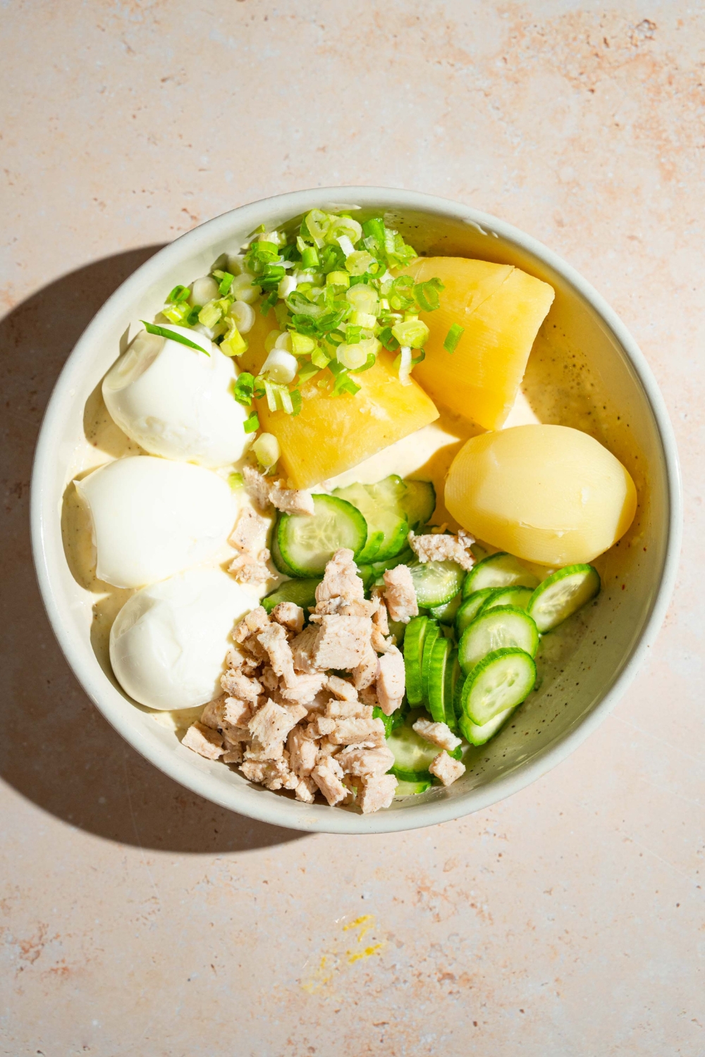 A large bowl with ingredients to make Japanese potato salad including cooked potatoes, cucumber, hard boiled eggs, chicken, and sliced green onion. The bowl is on a tan counter.