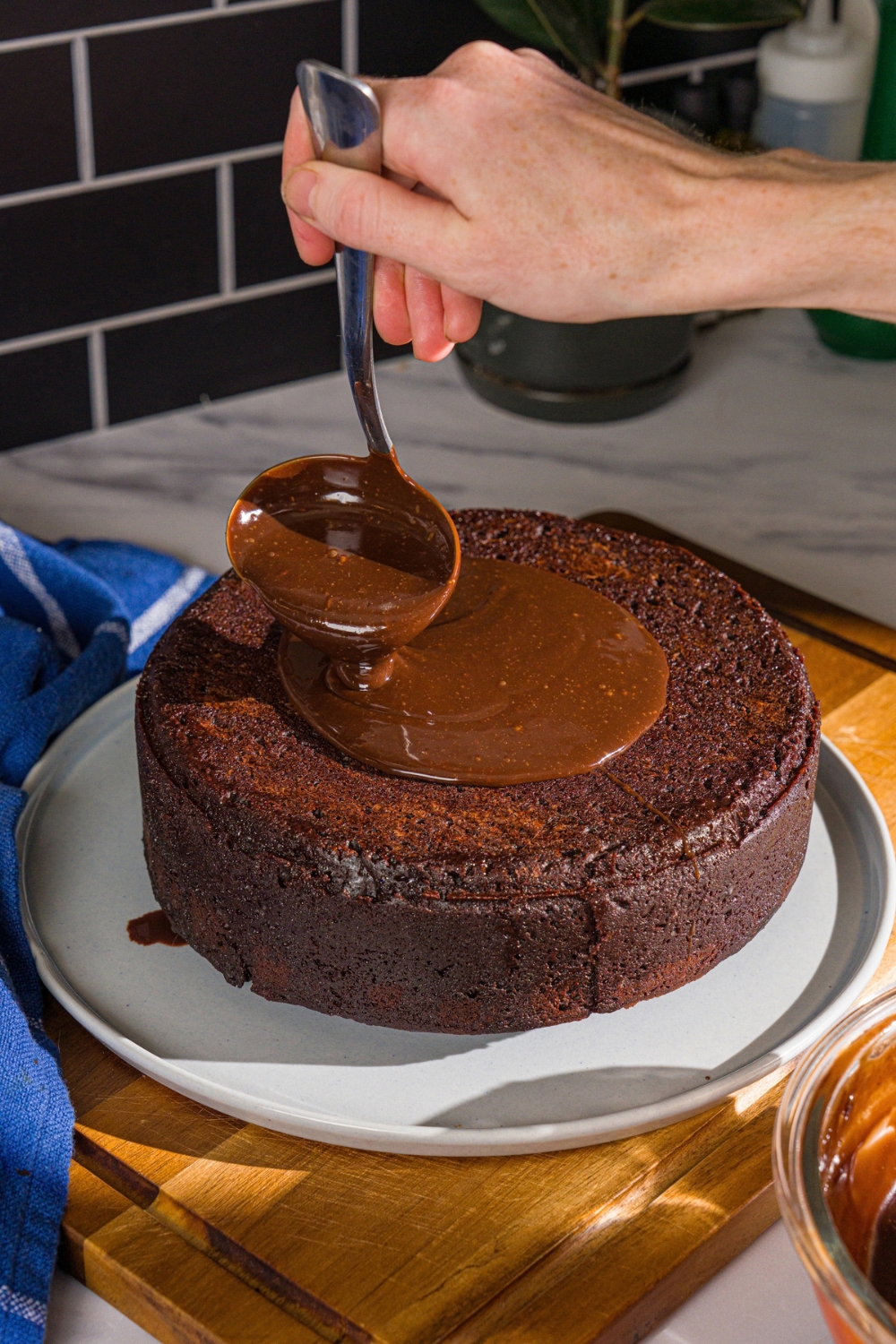A plate with a Matilda cake turned out onto the plate. A ladle is pouring chocolate ganache over the cake. The plate is on a wooden board.