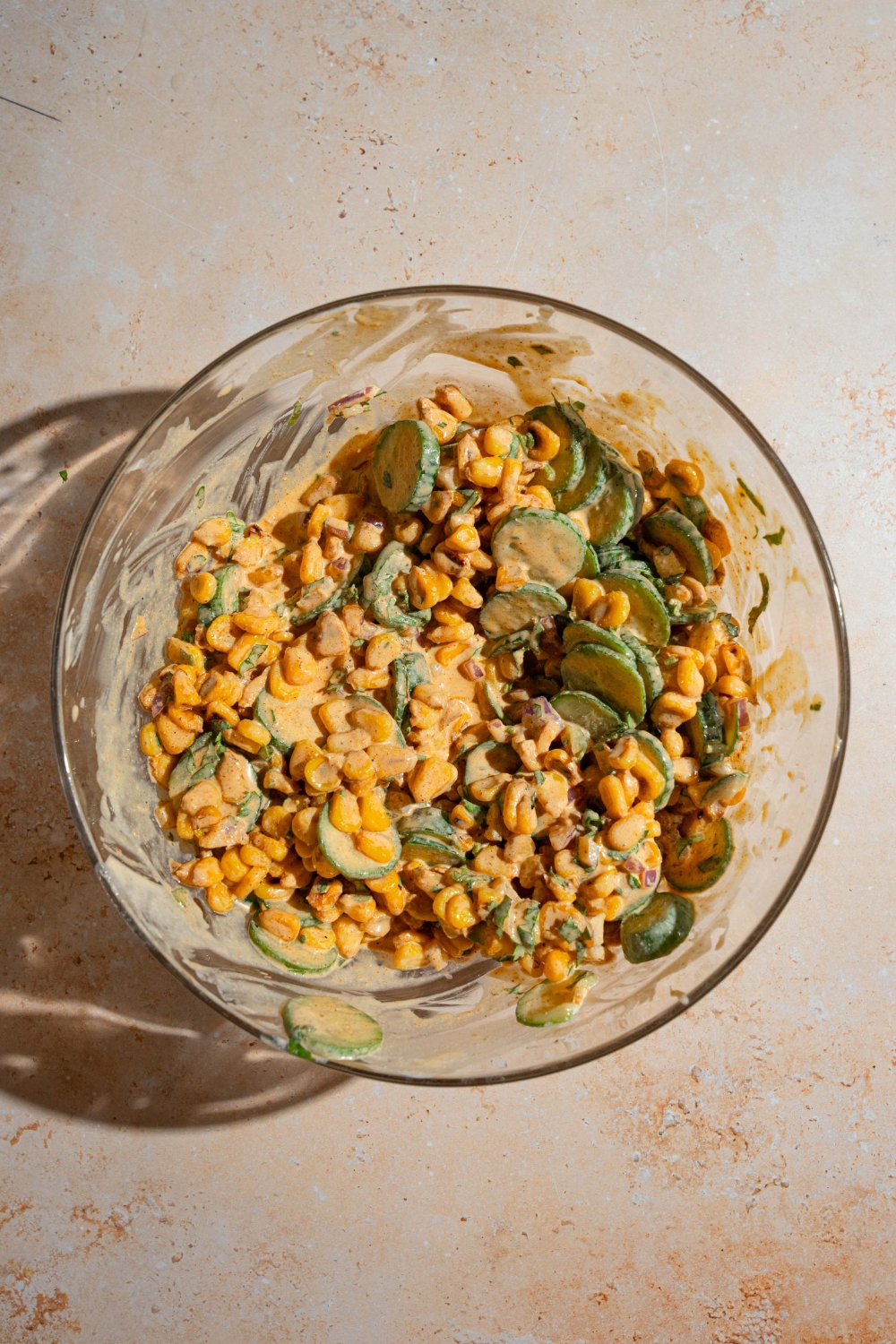 A large bowl of Mexican cucumber salad with corn, onion, and cilantro tossed in elote dressing. The bowl is on a tan counter.