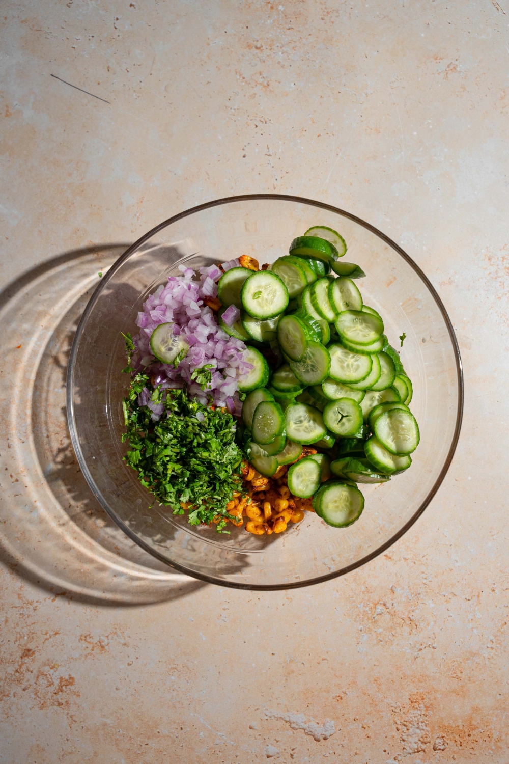 A large glass bowl with seasoned corn, sliced cucumbers, red onion, and cilantro. The bowl is on a tan counter.