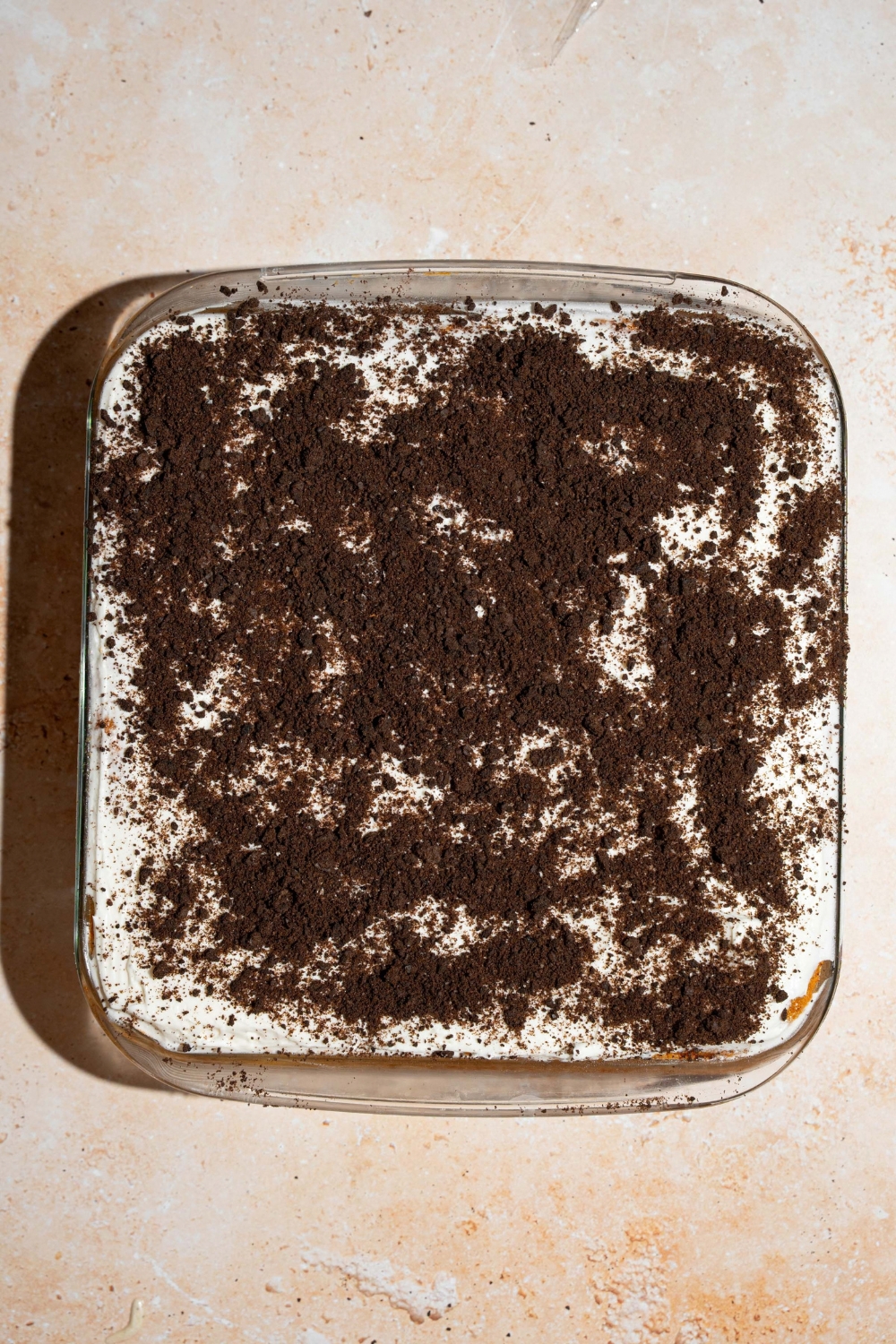 A square baking dish with baked Oreo tres leches cake topped with whipped cream and Oreo crumbles. The dish is on a tan counter.