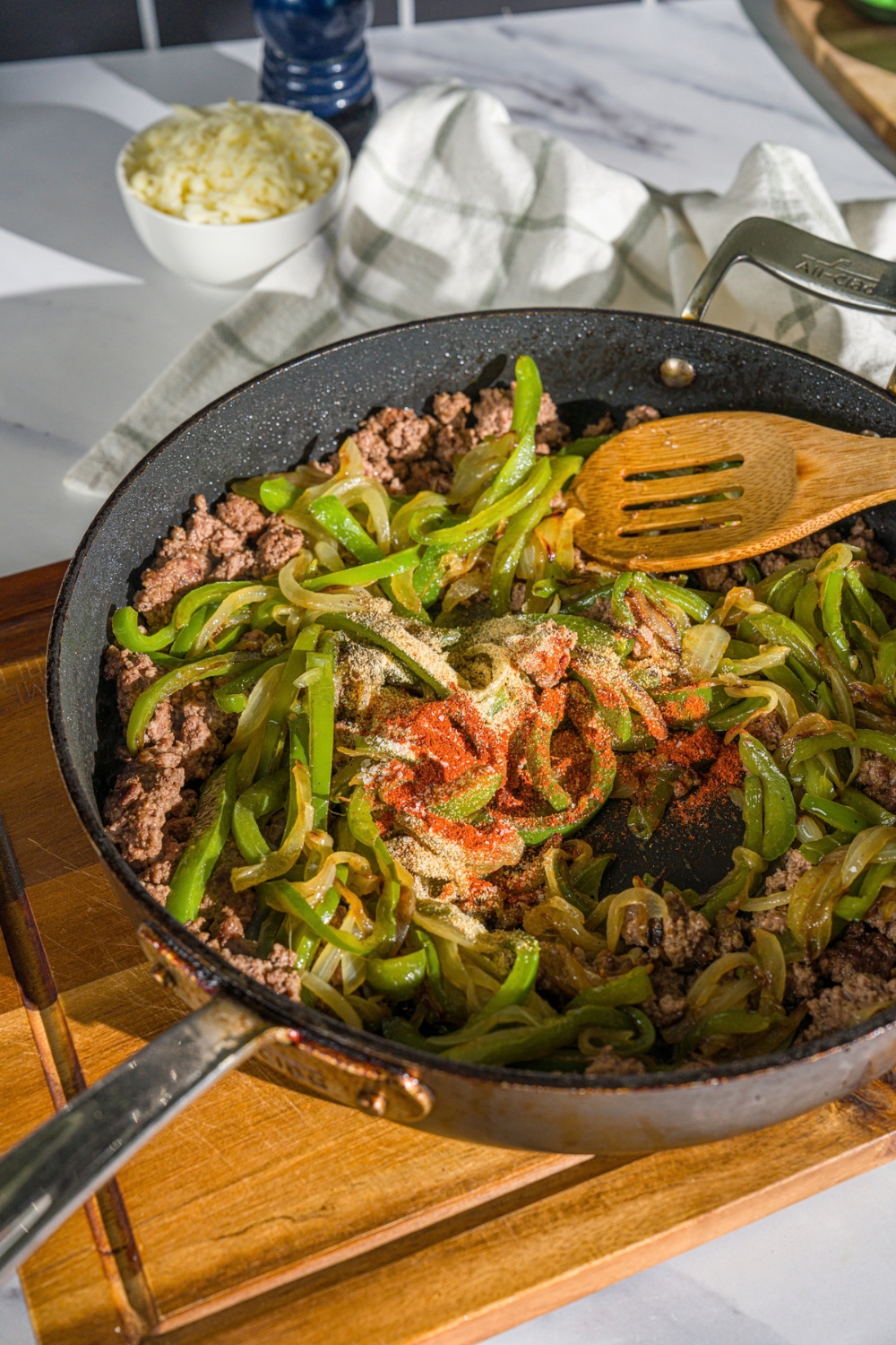 A skillet with cooked ground beef, sliced green peppers, and onions topped with seasonings. The skillet is on a wooden board.