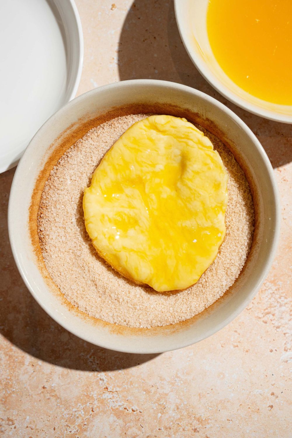 A small bowl with a cinnamon sugar mixture with a biscuit dipped in egg. The bowl is on a tan counter.