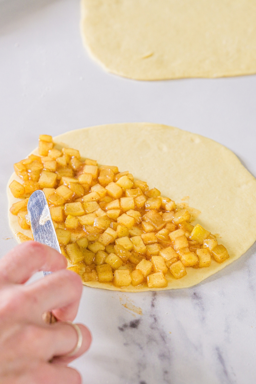 Apple pie filling being spread over the bottom half of a round piece of dough. The dough is on a marble counter.