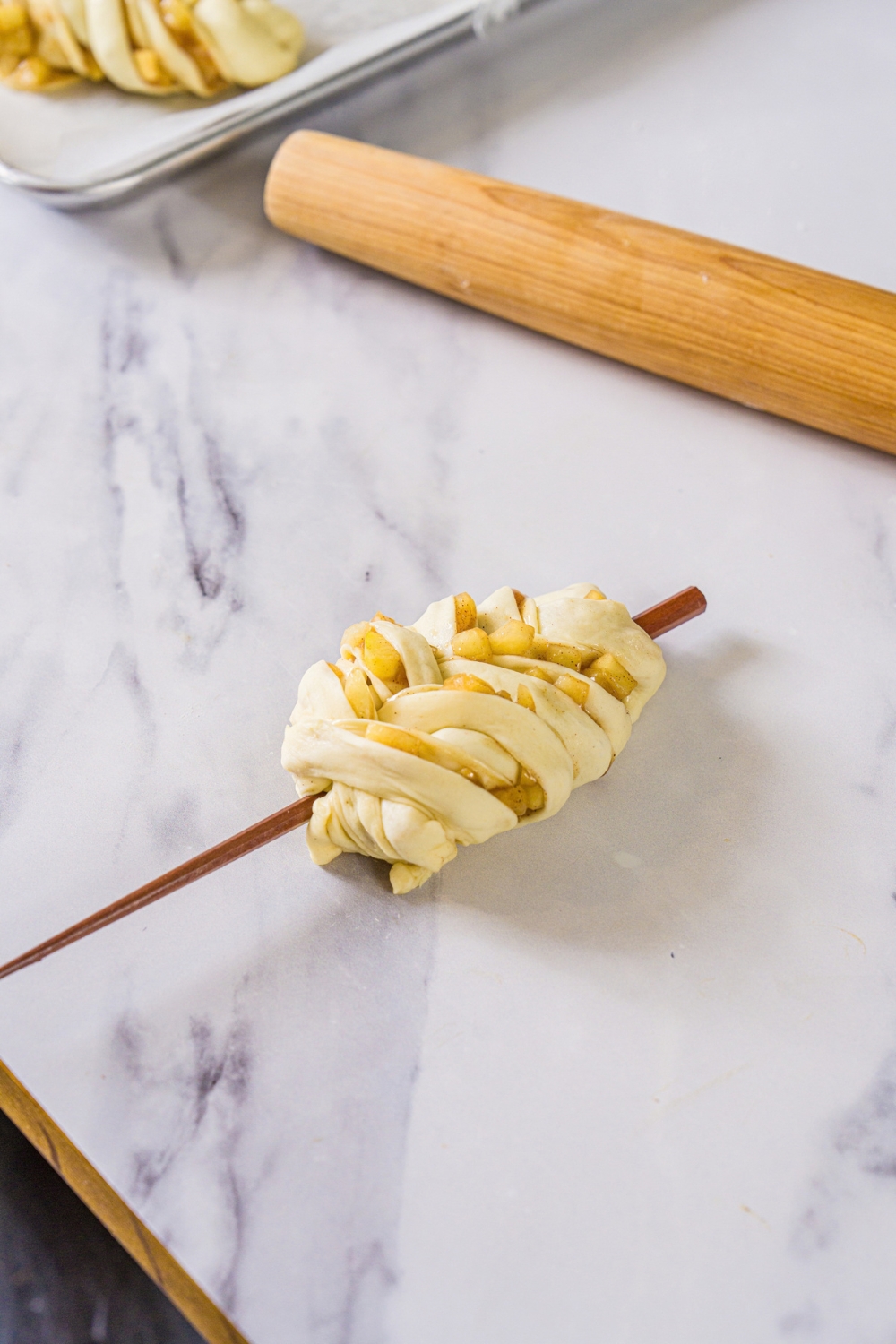 An unbaked apple pie roll twisted and braided around a chopstick. The roll is on a marble counter.