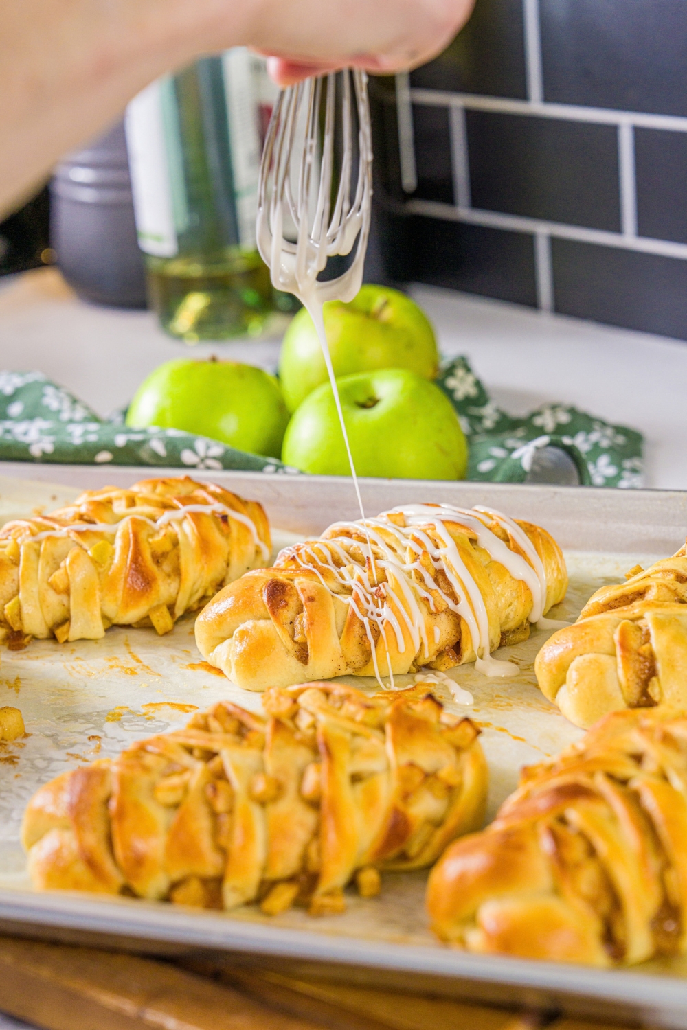 A baking sheet lined with parchment paper with baked twisted apple pie rolls. A whisk is drizzling icing over the rolls.