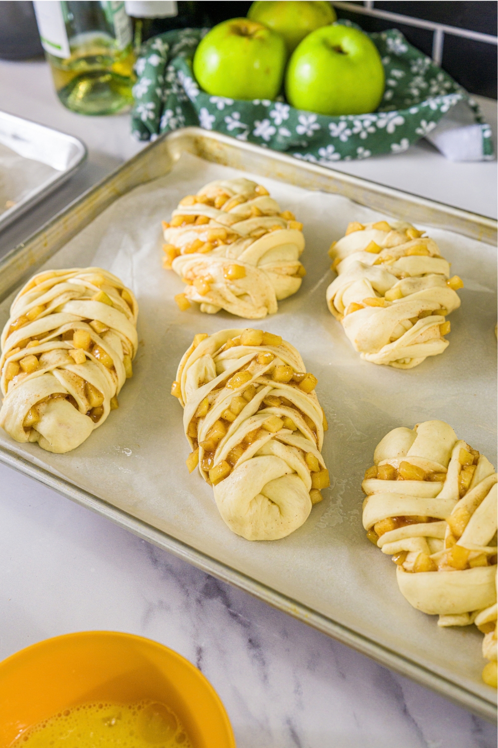 A baking sheet lined with parchment paper with unbaked twisted apple pie rolls. The sheet is on a marble counter.
