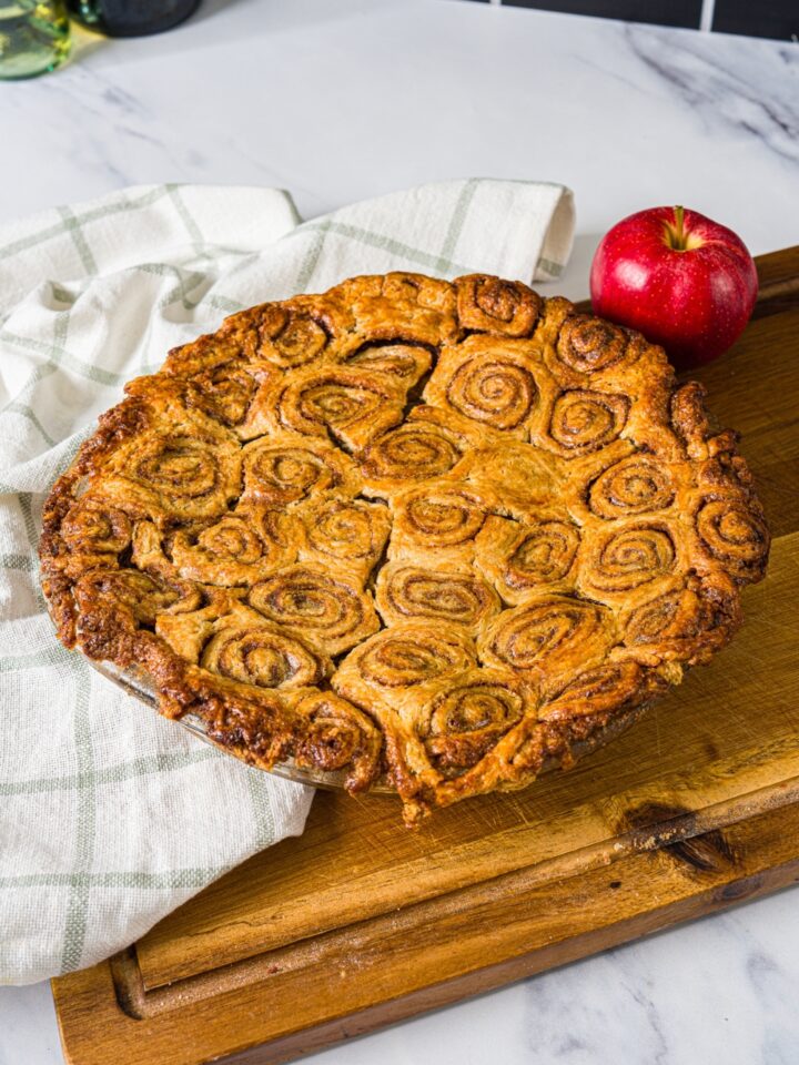 A wooden board with a glass pie dish with baked cinnamon roll apple pie. The board is on a marble counter with a white cloth napkin.