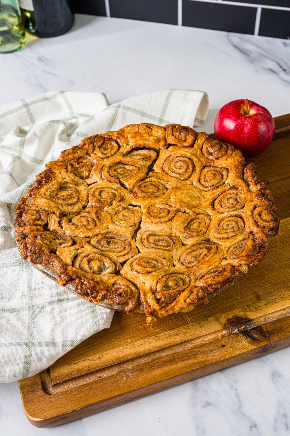 A wooden board with a glass pie dish with baked cinnamon roll apple pie. The board is on a marble counter with a white cloth napkin.
