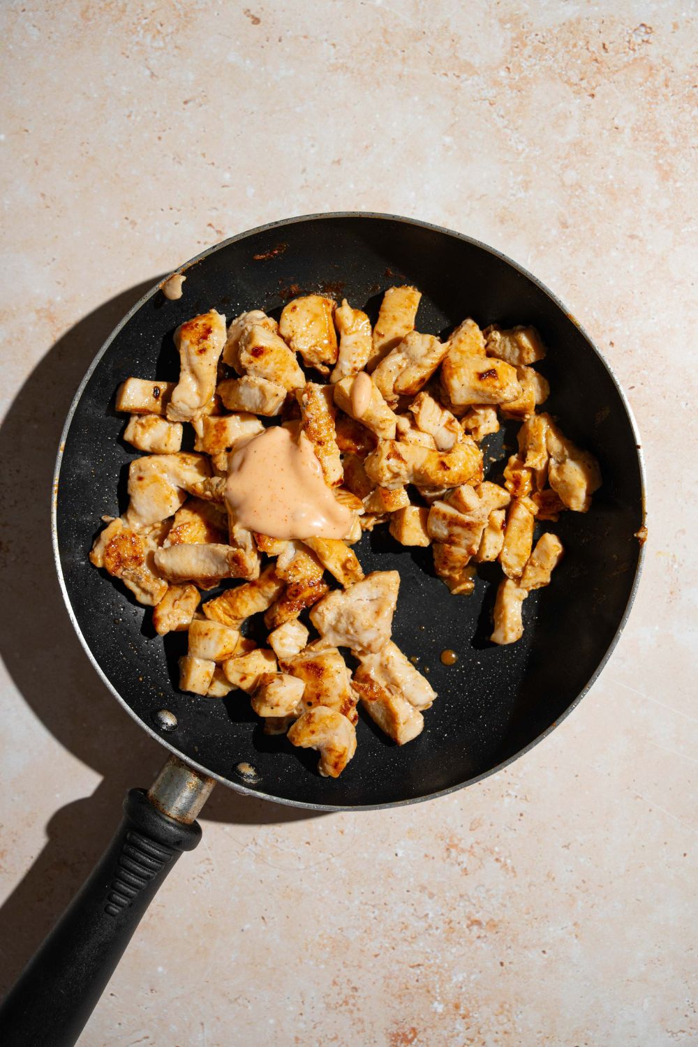 A skillet with seared chicken pieces topped with bang bang sauce. The skillet is on a tan counter.