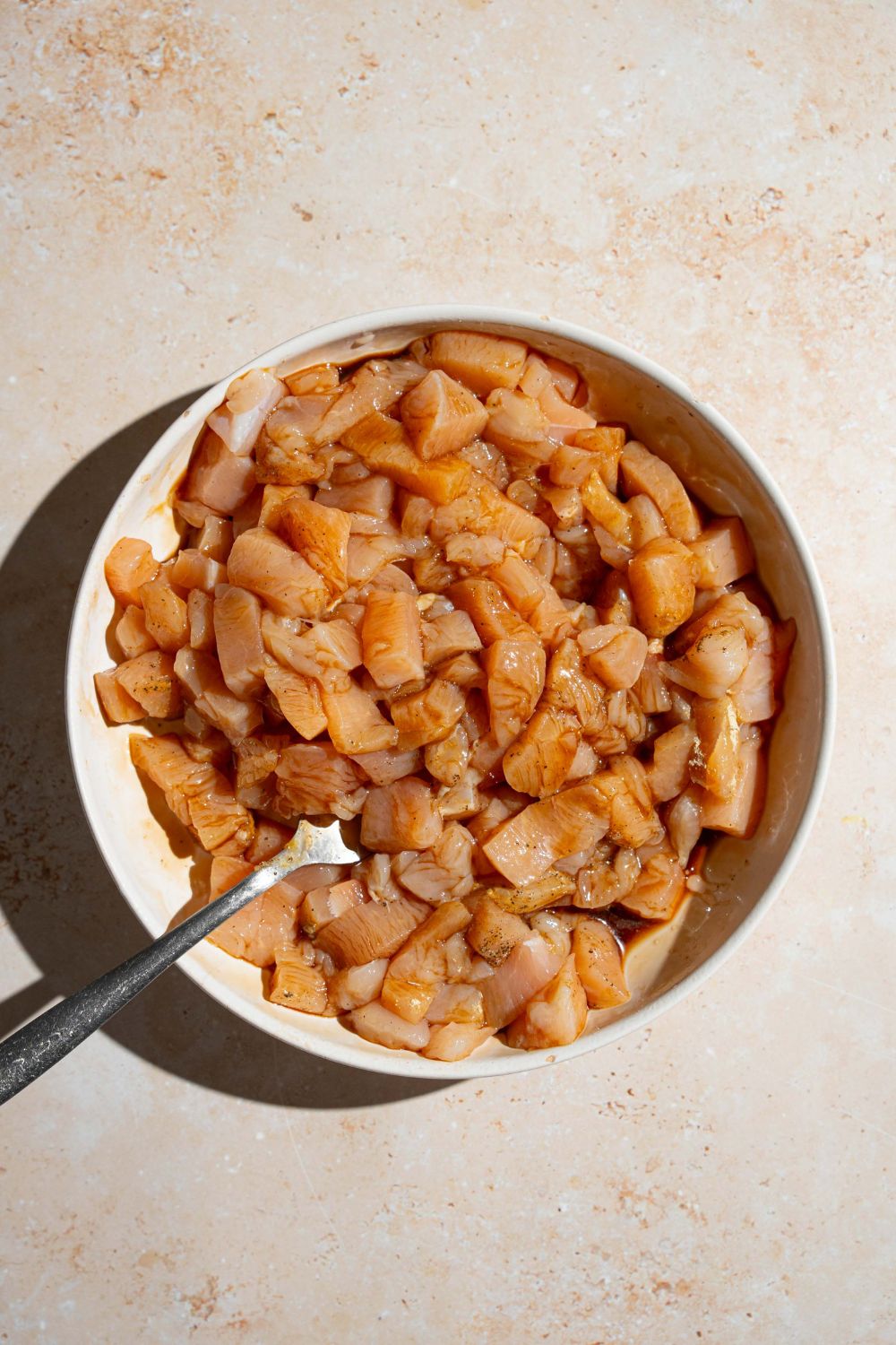 A large white bowl with cubed chicken tossed in seasonings. There is a fork in the bowl. The bowl is on a tan counter.