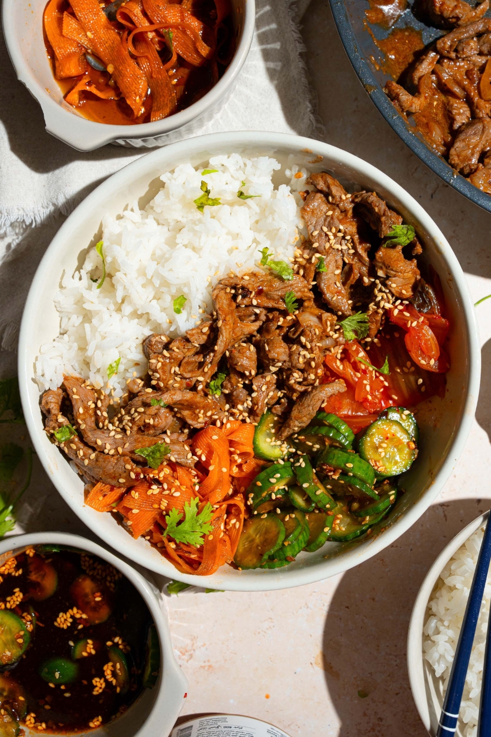 A white bowl with a beef bulgogi bowl served with white rice, beef bulgogi, and a cucumber carrot salad, all garnished with sesame seeds. The bowl is on a tan counter with bowls of ingredients.