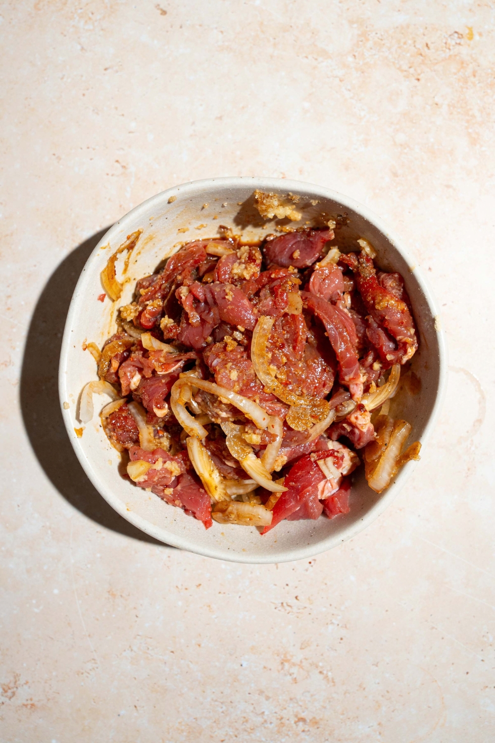 A white bowl with uncooked beef strips in tossed in marinade with white onions. The bowl is on a tan counter.