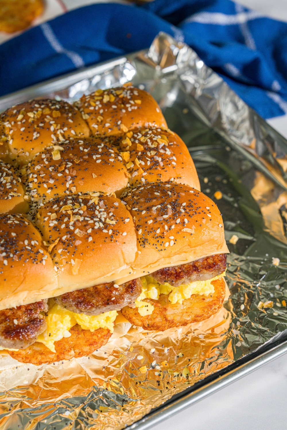 A baking sheet lined with foil with baked wooden board with breakfast sliders made with sausage, hashbrowns, scrambled eggs, and cheese on seasoned hawaiian rolls.