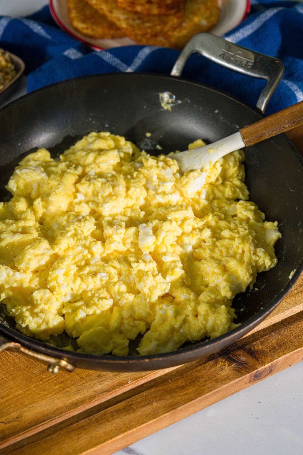 A skillet with a spatula stirring scrambled eggs. The skillet is on a wooden board.