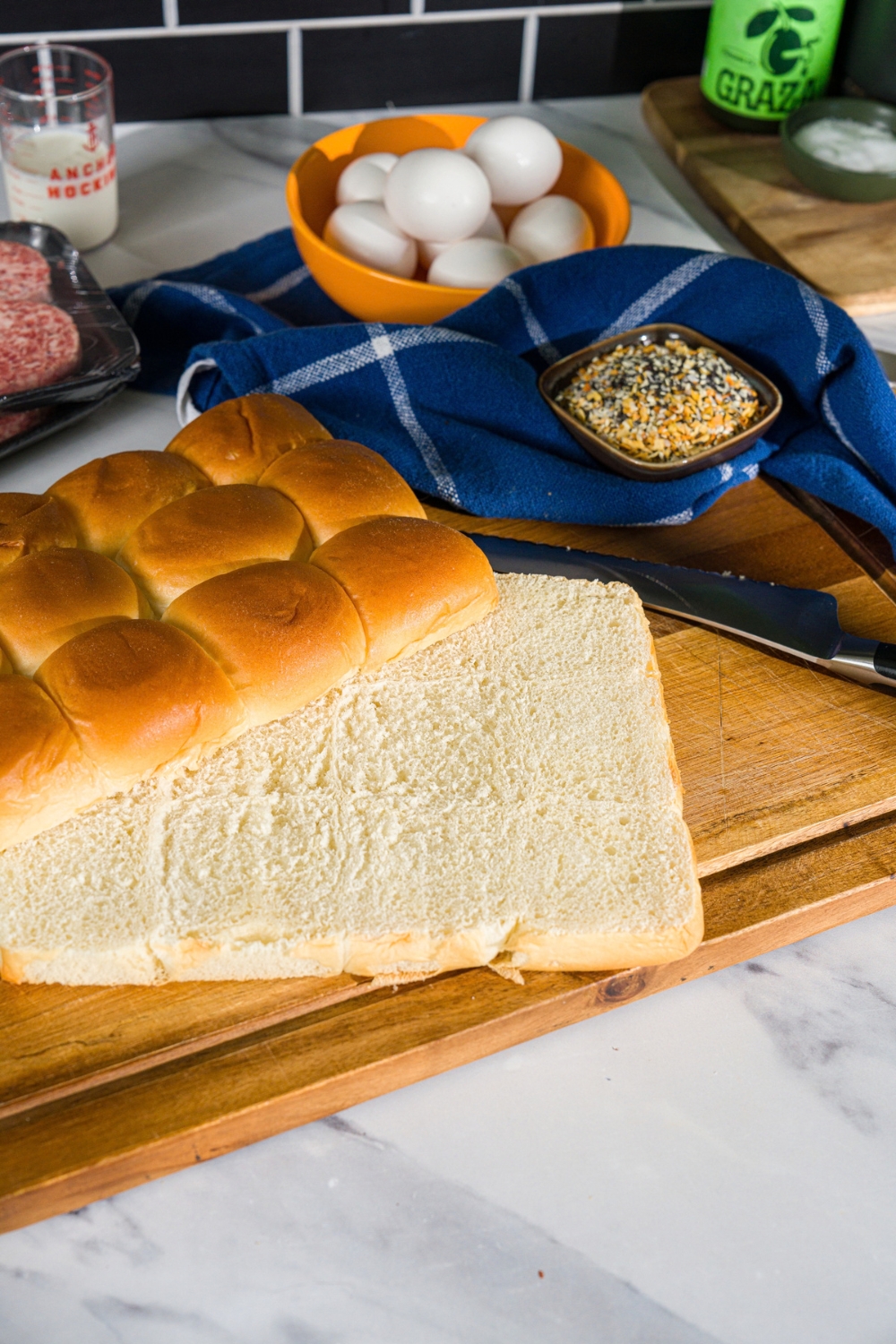 A wooden board with an array of hawaiian rolls sliced in half with a bowl of seasonings and blue cloth napkin.