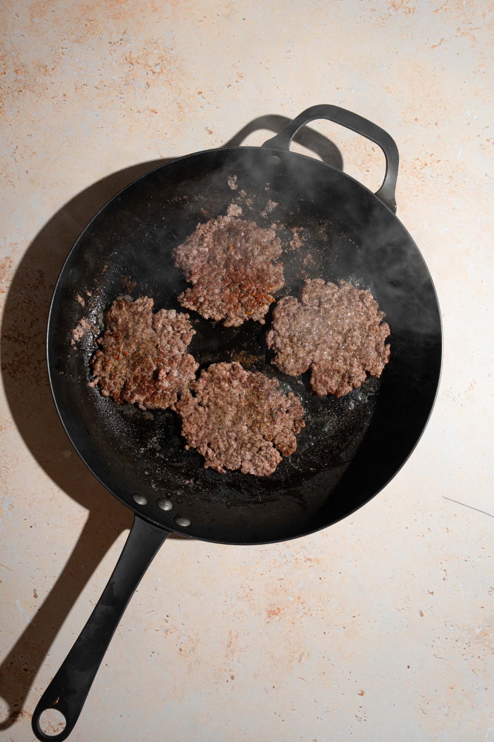 A skillet with four smashed burger patties cooking on the skillet. The skillet is on a tan counter.