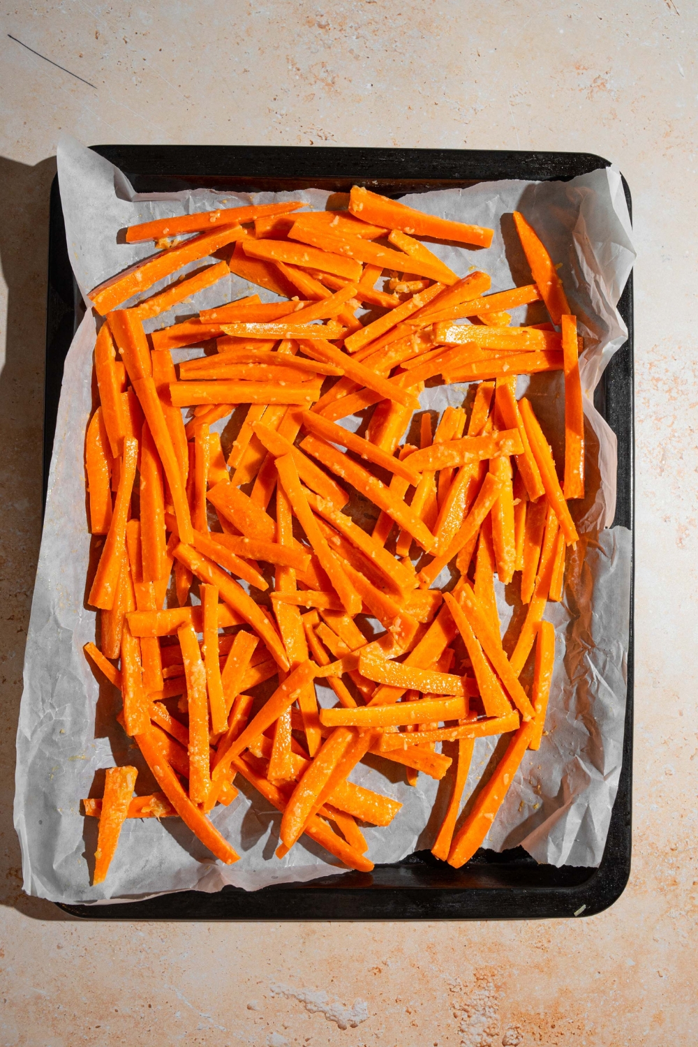 A baking sheet lined with parchment paper with unbaked seasoned carrot fries. The sheet is on a tan counter.