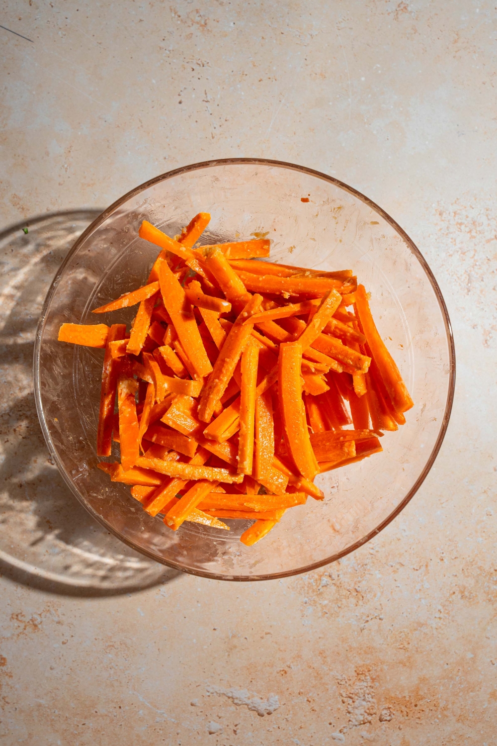 A bowl of sliced carrots tossed in a seasoning mixture. The bowl is on a tan counter.
