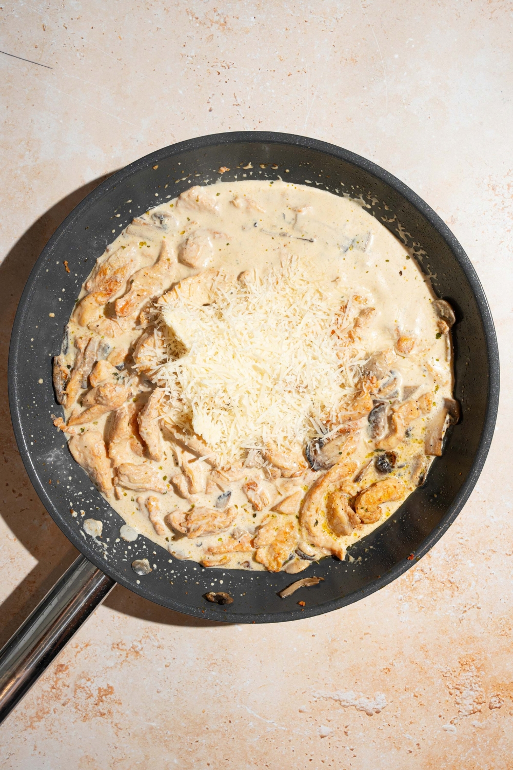 A skillet with chicken mushroom alfredo sauce base topped with parmesan cheese. The skillet is on a tan counter.