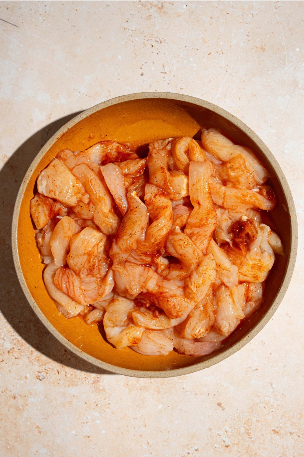 A bowl of uncooked chicken strips tossed in seasonings. The skillet is on a tan counter.