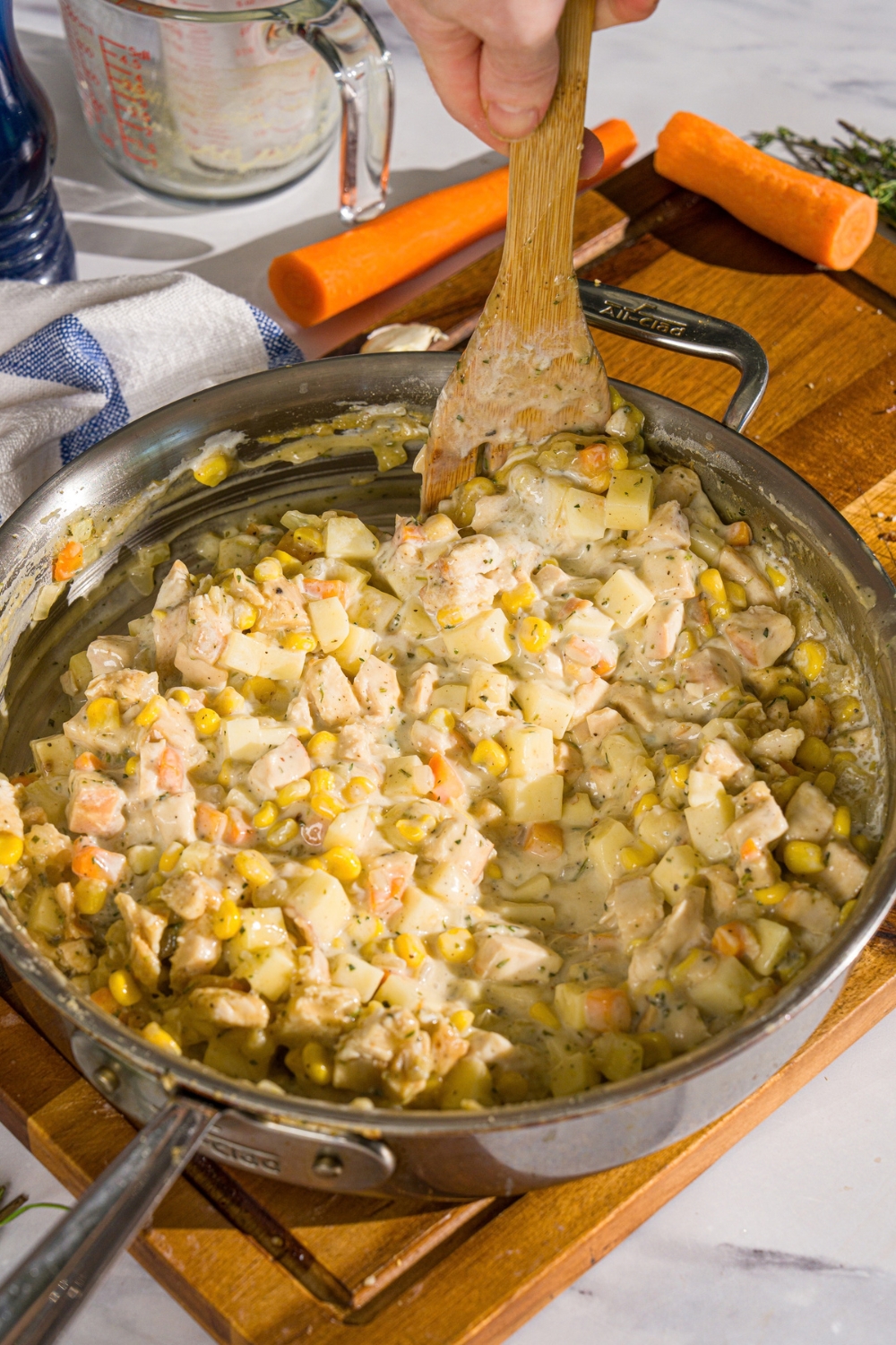 A skillet with a wooden spoon mixing chicken pot pie filling. The skillet is on a wooden board.
