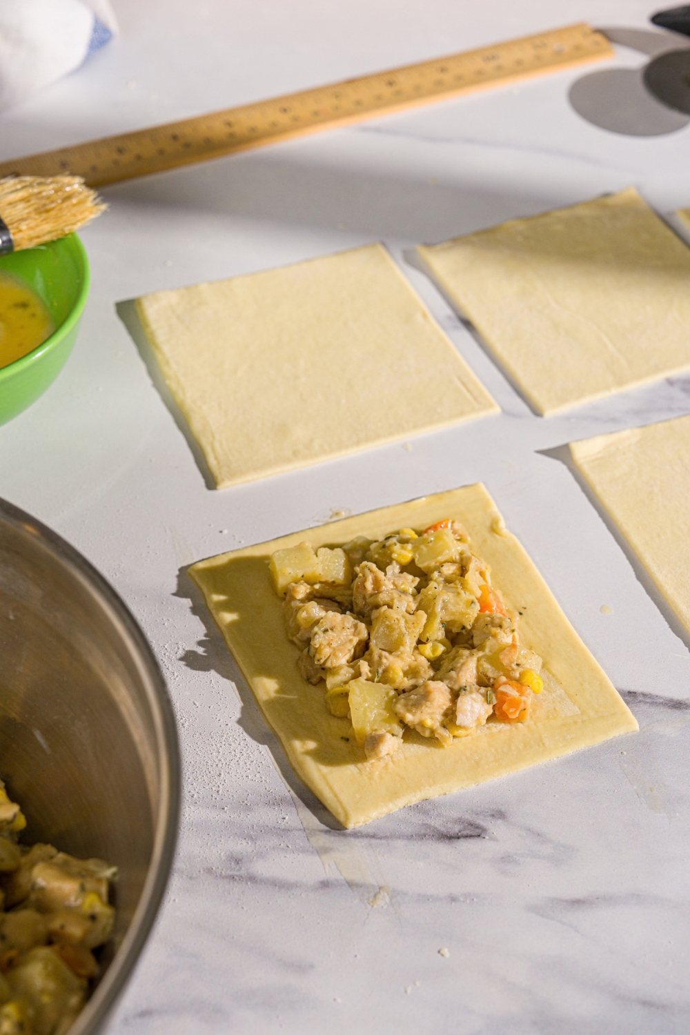 A marble counter with puff pastry sheets sliced into squares with chicken pot pie filling in the center of one of the pastries.