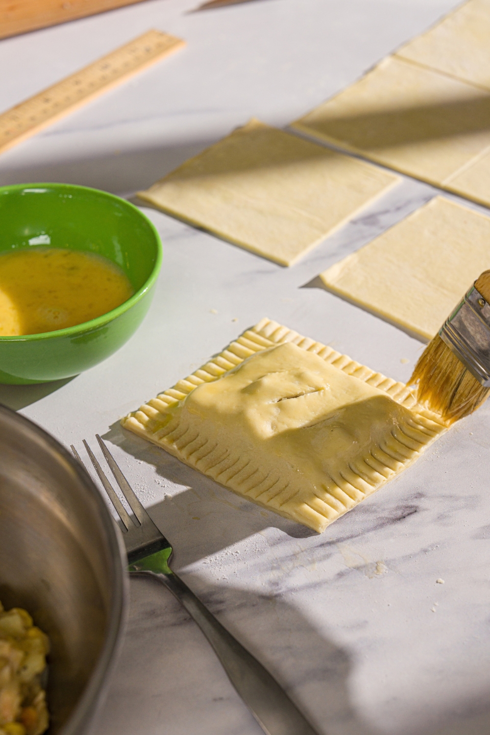 A sealed, unbaked chicken pot pie hand pie being brushed with egg wash. The hand pie is on a marble counter with additional puff pastry square and a bowl of egg wash.