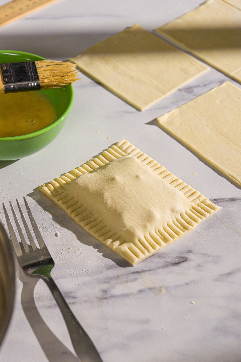 A sealed, unbaked chicken pot pie hand pie. The hand pie is on a marble counter with additional puff pastry squares and a bowl of egg wash.