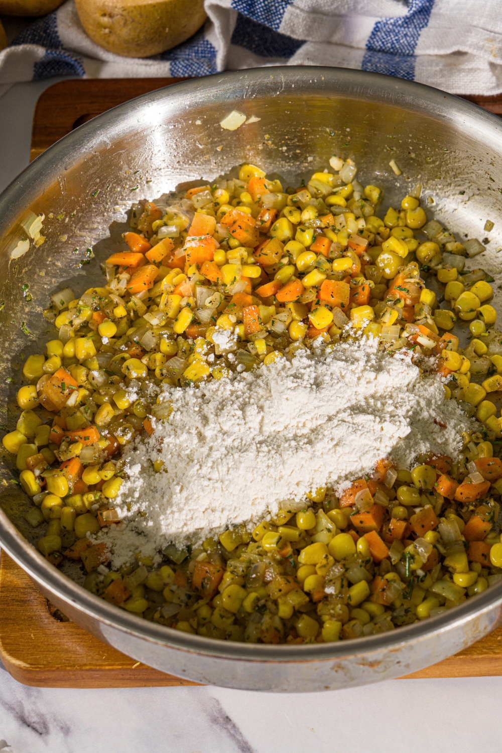 A skillet with sautéed corn, carrots, and onions with flour on top. The skillet is on a wooden board.