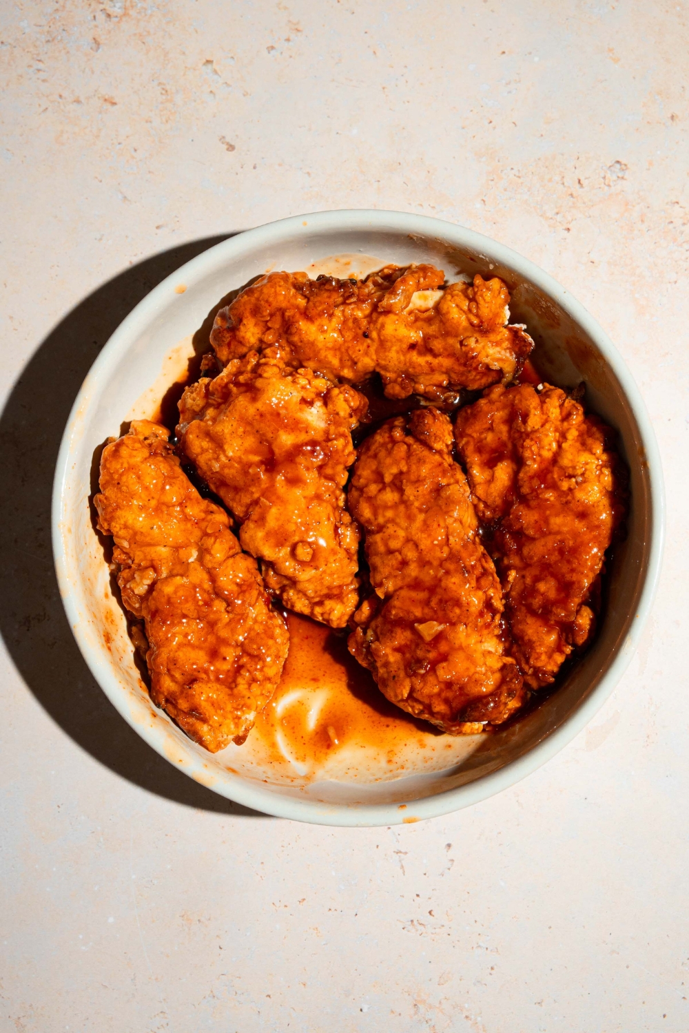 A large white bowl with fried chicken tenders tossed in sweet chili sauce. The bowl is on a tan counter.