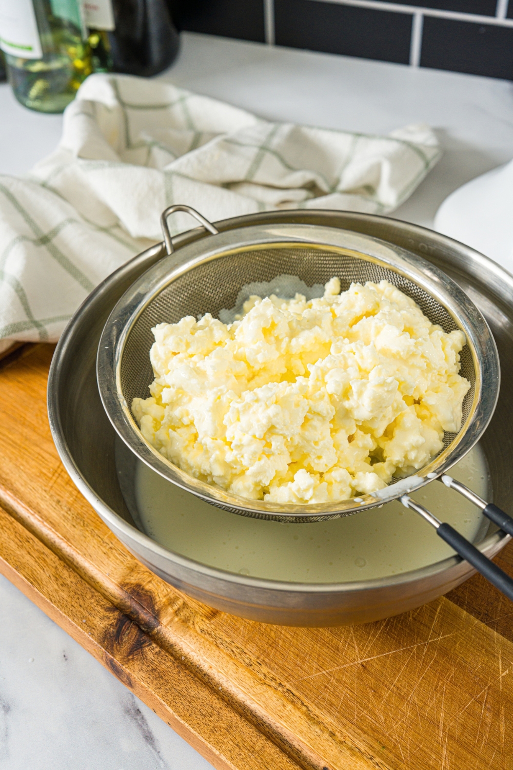 A cultured butter mixture being strained through a strainer over a large bowl. The bowl is on a wooden board.