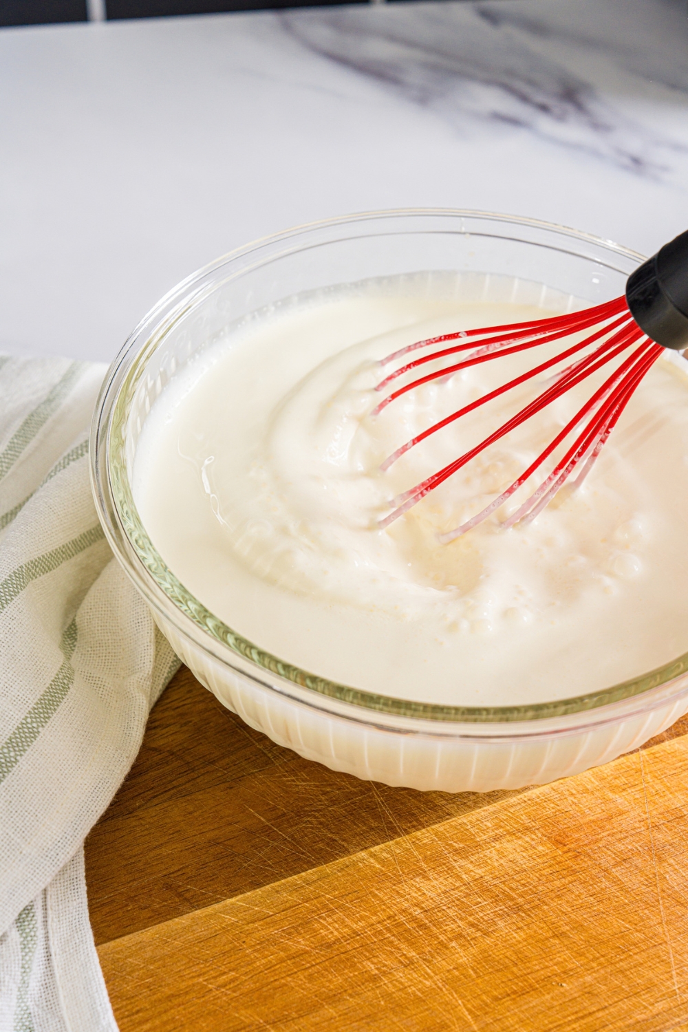 A large bowl with a whisk mixing heavy cream and yogurt. The bowl is on a wooden board.