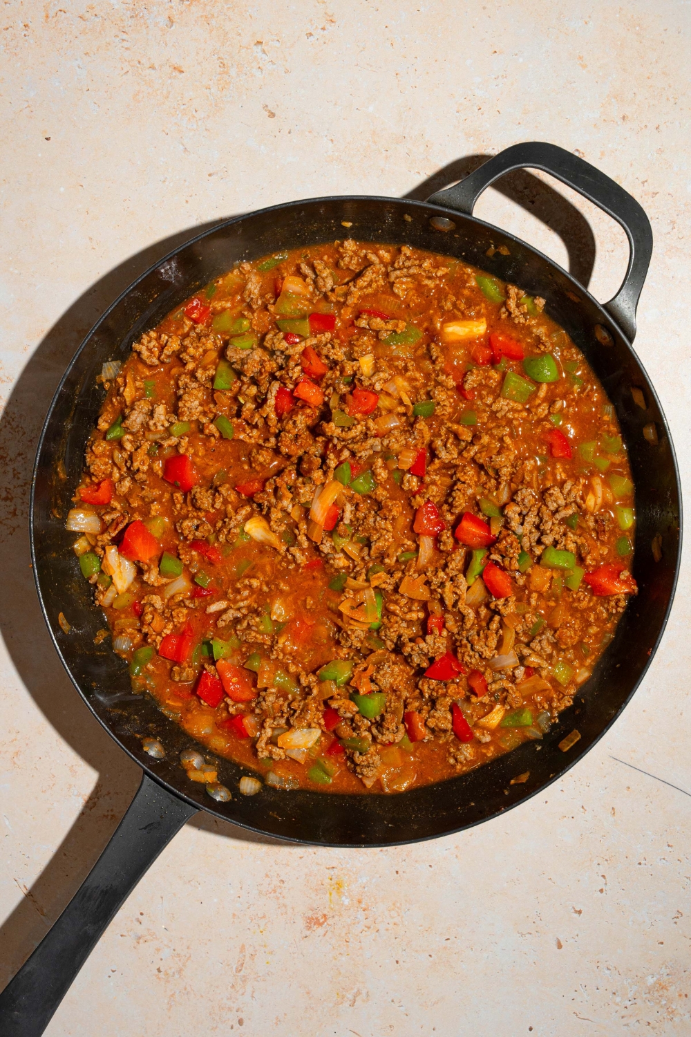 A skillet with dirty spaghetti sauce mixture including ground beef, sausage, diced vegetables, tomato paste, beef stock, and more. The skillet is on a tan counter.