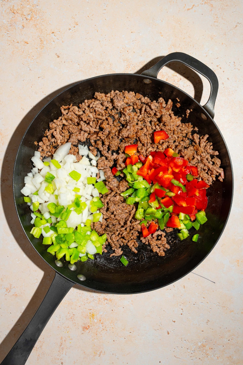 A skillet with cooked ground beef and Italian sausage with diced bell peppers, celery, and onion. The skillet is on a tan counter.