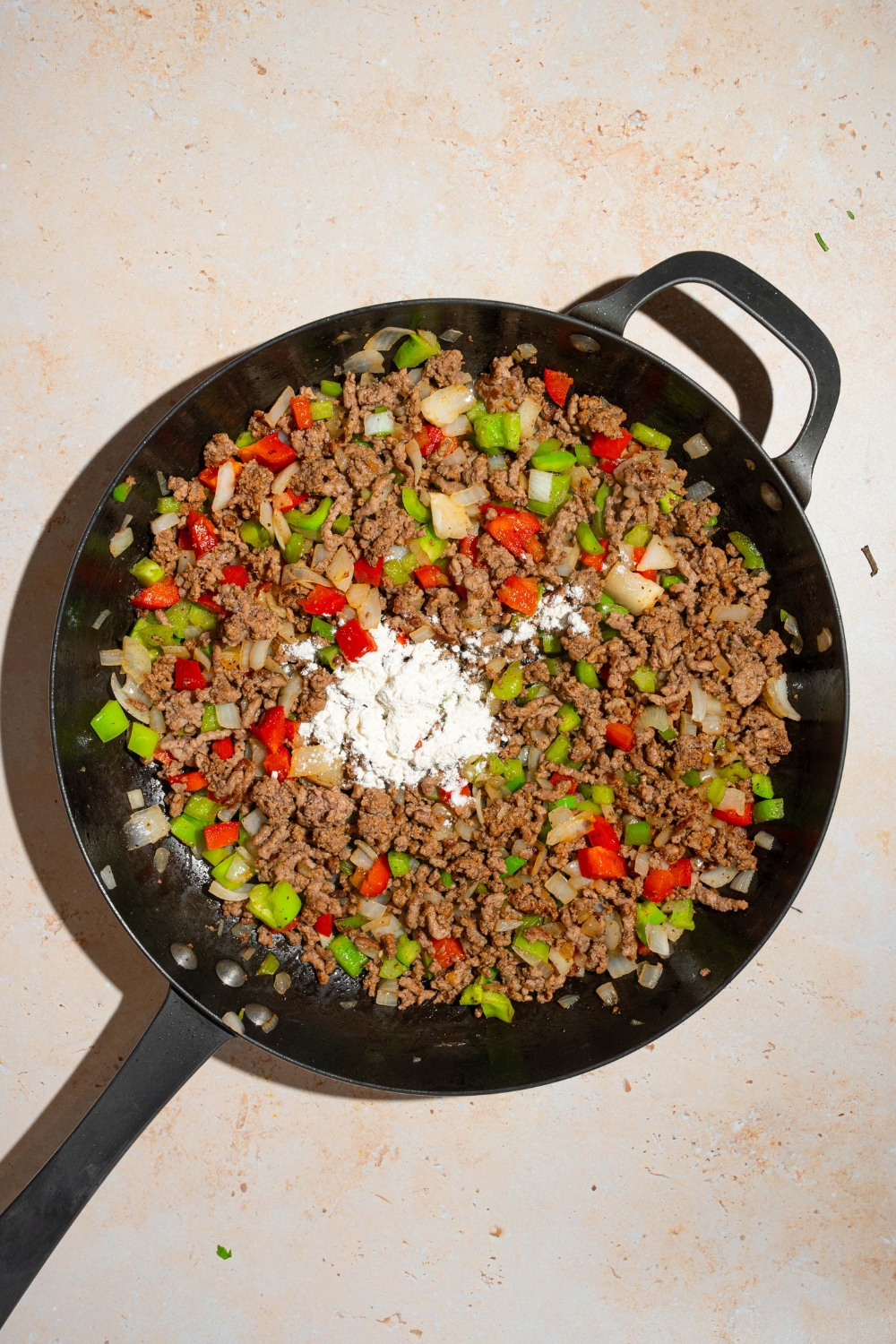 A skillet with cooked ground beef, Italian sausage, and diced vegetables, topped with flour. The skillet is on a tan counter.