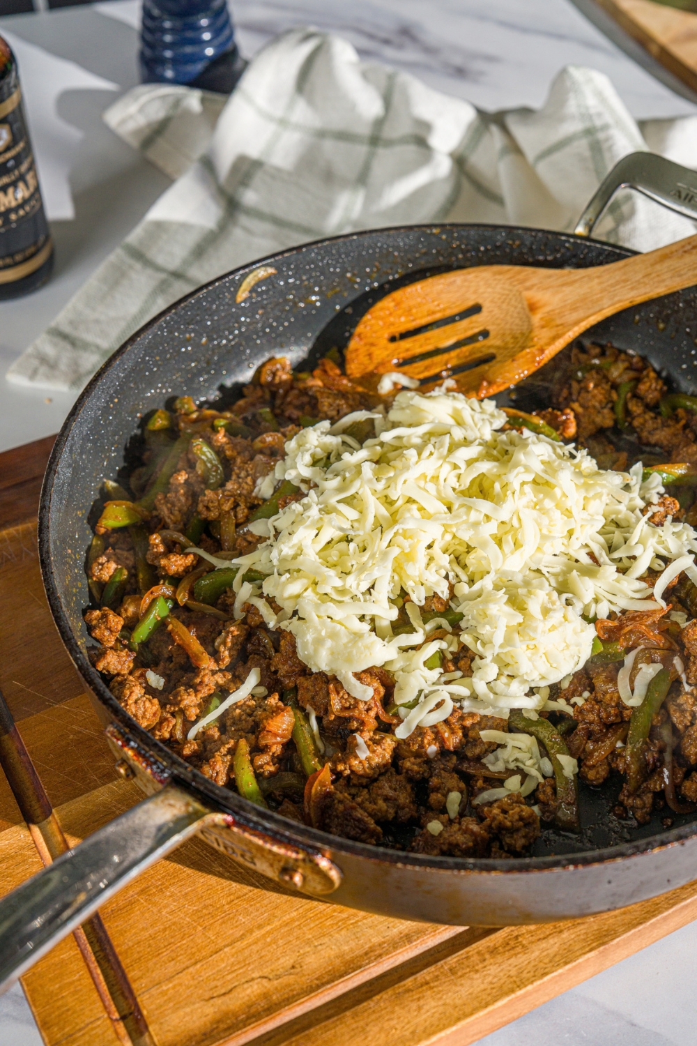 A skillet with a wooden spoon stirring a ground beef, green peppers, onion, and seasonings topped with shredded cheese. The skillet is on a wooden board.