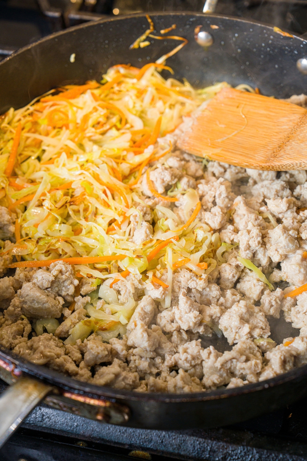 A skillet with sautéed cabbage and carrots with cooked ground chicken. There is a wooden spatula in the mixture. The skillet is cooking on a stove.
