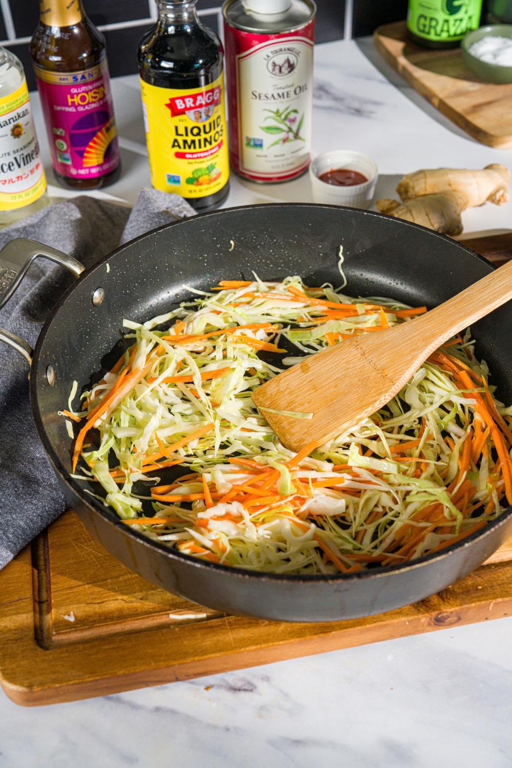 A skillet with a wooden spatula mixing sliced cabbage and carrots. The skillet is on a wooden board on a marble counter.