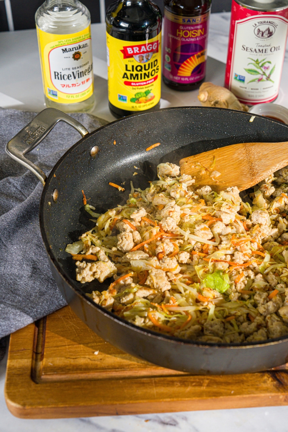 A skillet with a wooden spatula combining cooked ground chicken with sautéed cabbage and carrots and seasonings. The skillet is on a wooden board with a blue cloth napkin.