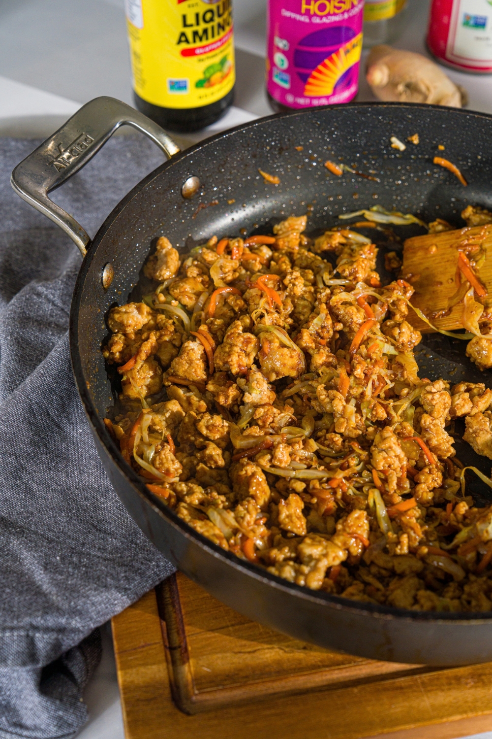 A skillet with ground chicken and sautéed cabbage and carrots tossed in an egg roll sauce. The skillet is on a wooden board with a blue cloth napkin.