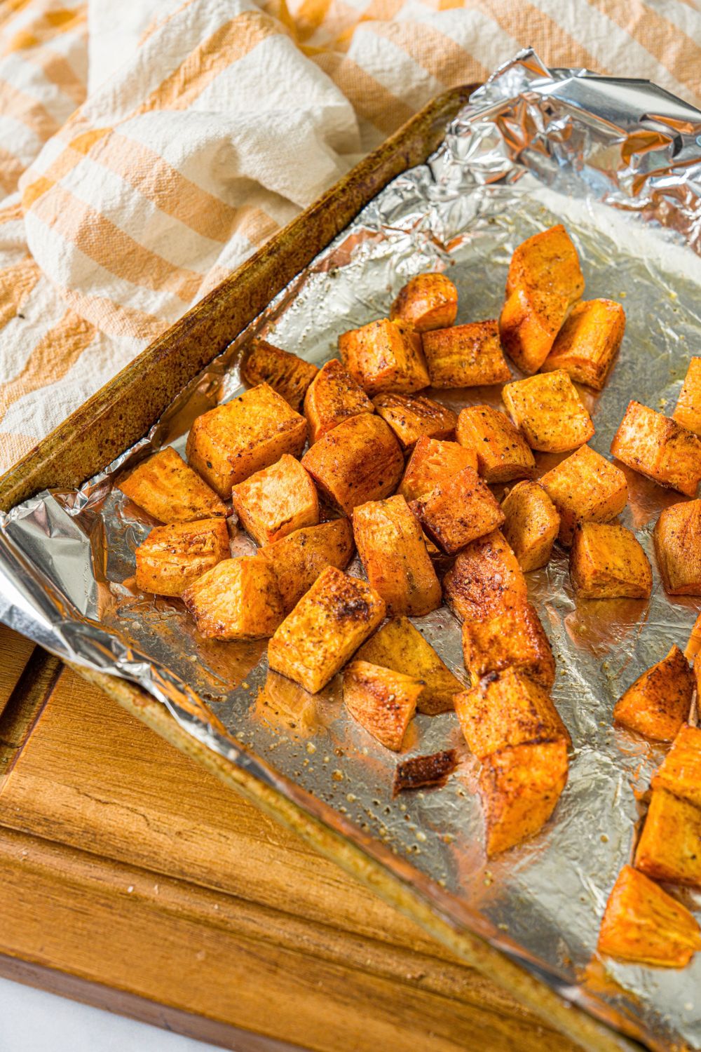 A sheet pan lined with foil with roasted sweet potatoes. The pan is on a wooden board with a yellow checkered napkin.