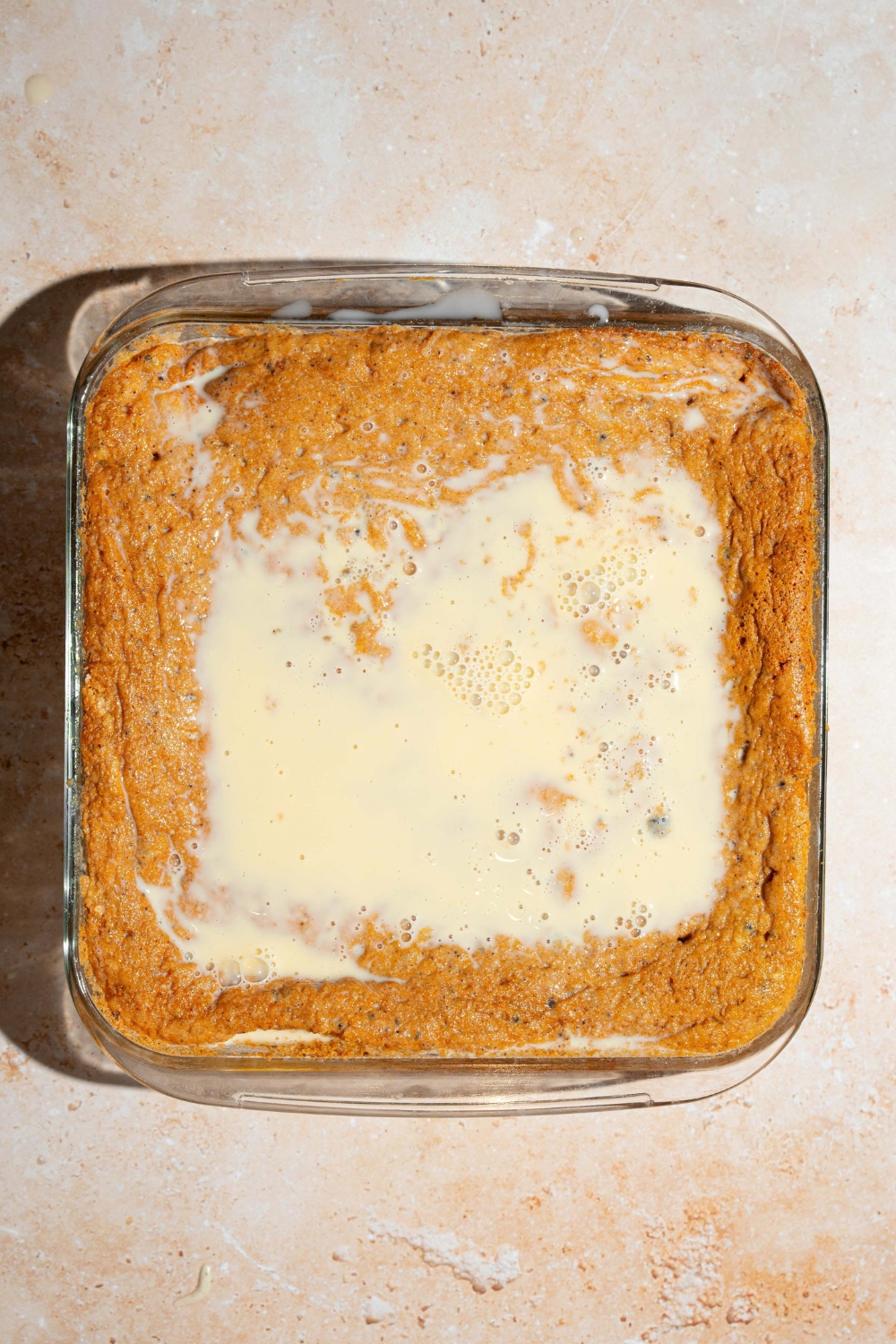 A square baking dish with baked Oreo tres leches cake topped with tres leches soak. The dish is on a tan counter.