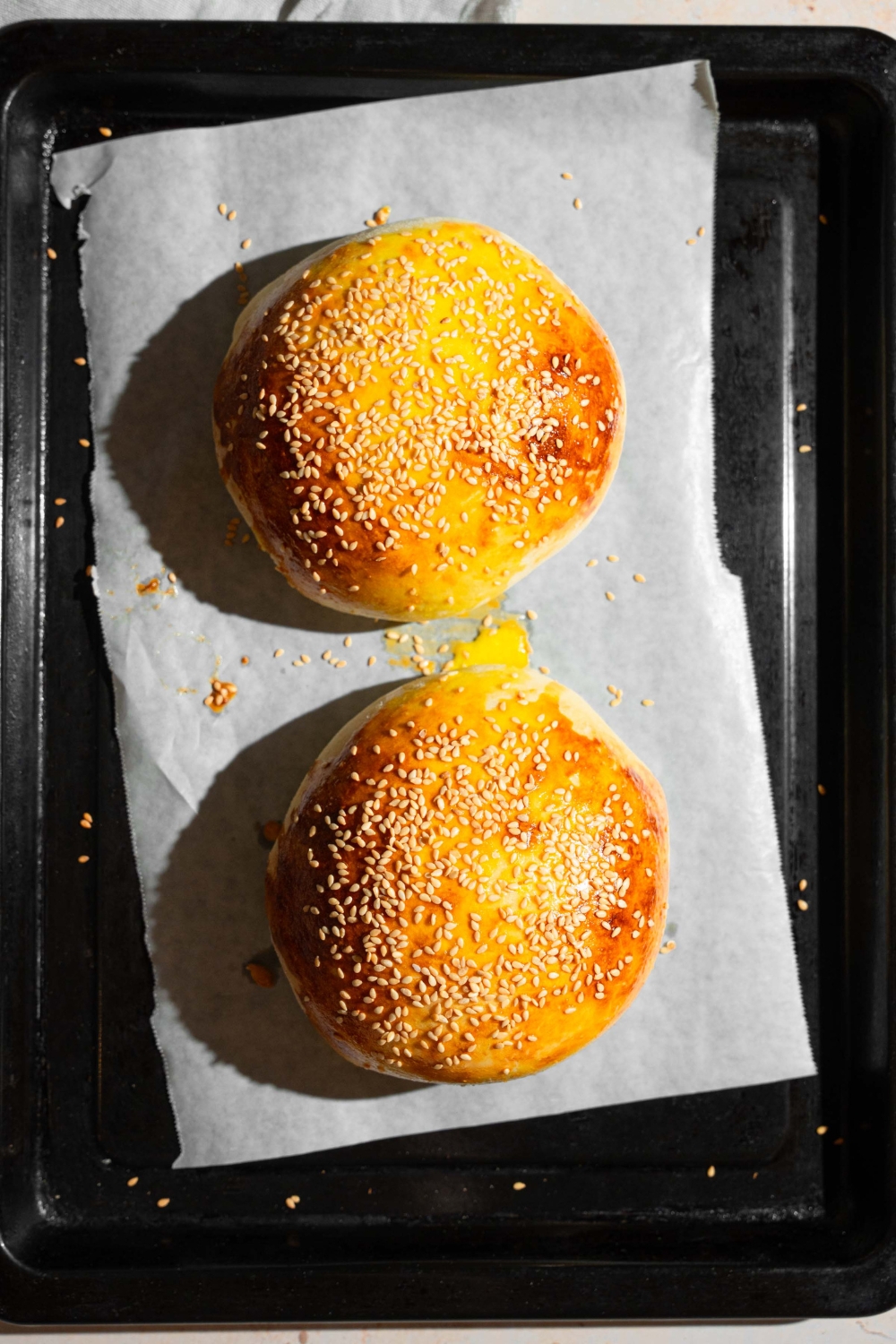 A baking sheet lined with parchment paper with two baked burger stuffed buns topped with sesame seeds.